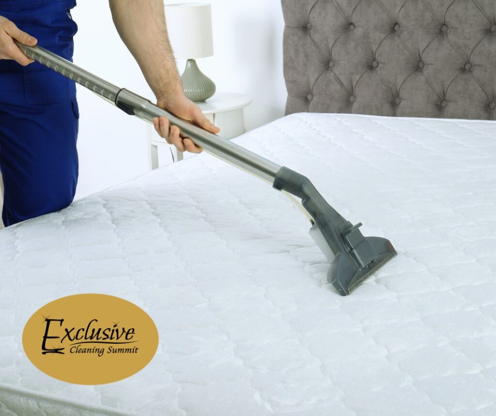 A person wearing a blue uniform with a vacuum performs a professional mattress cleaning. There is a gold and black logo for Exclusive Cleaning Summit a vacation rental cleaning company based in Frisco, Colorado.